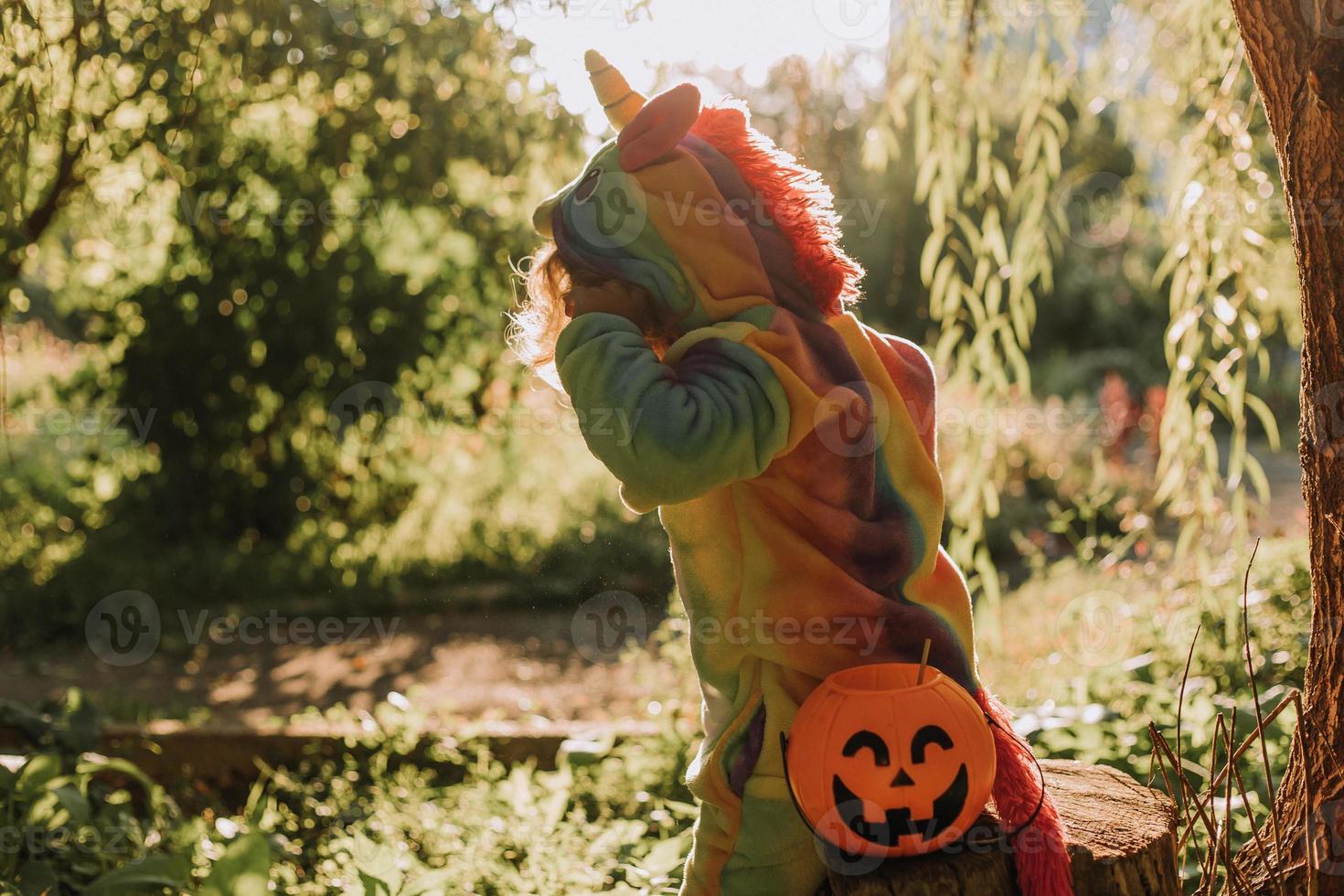 little girl in a rainbow unicorn Halloween costume and a dachshund in a dress with a pumpkin basket for sweets are sitting on stump at forest sunset. fabulous wonderful magical forest. space for text photo