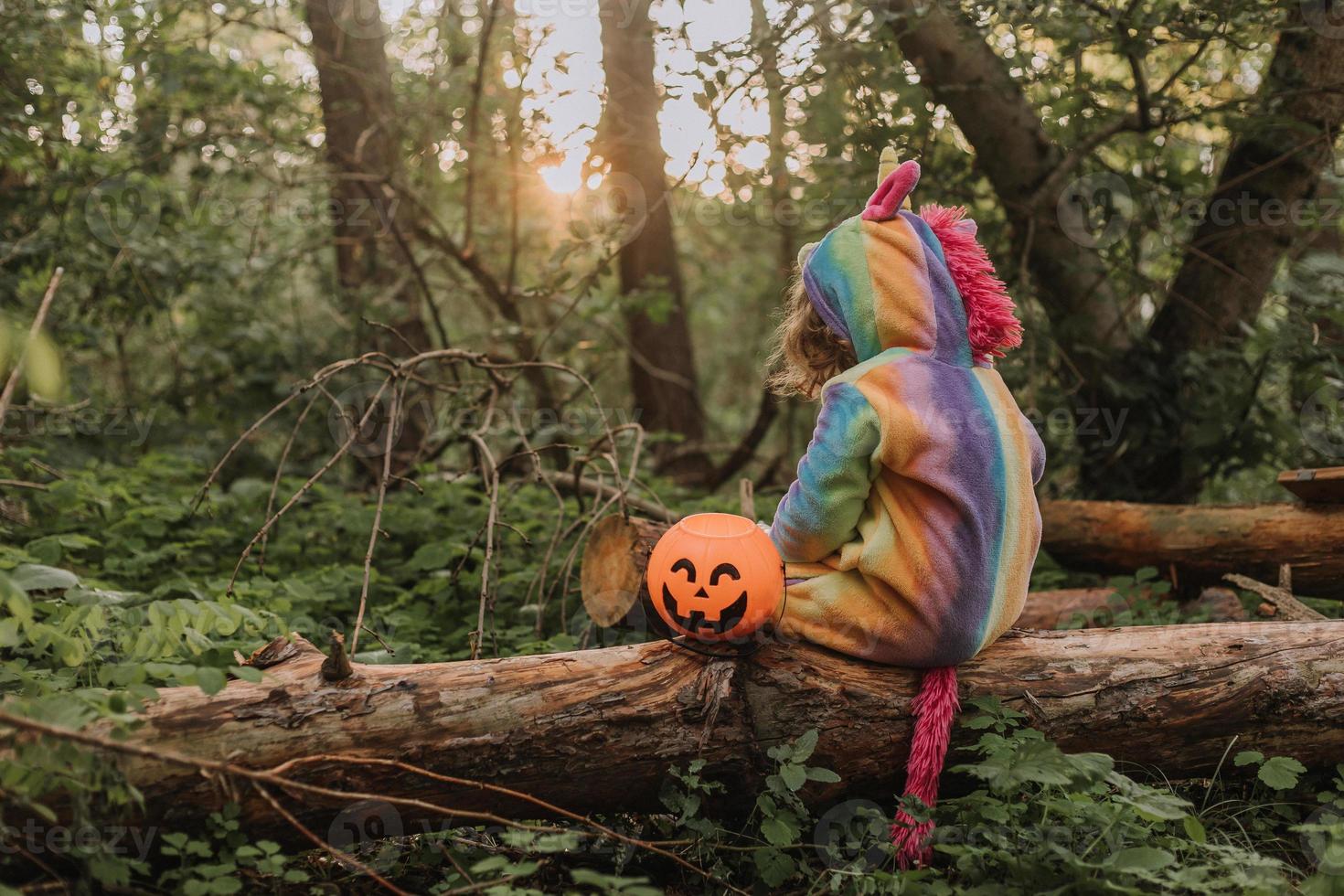 little girl in a rainbow unicorn Halloween costume with a pumpkin basket for sweets is sitting on a stump at a forest sunset. a fabulous wonderful magical forest. space for text. High quality photo