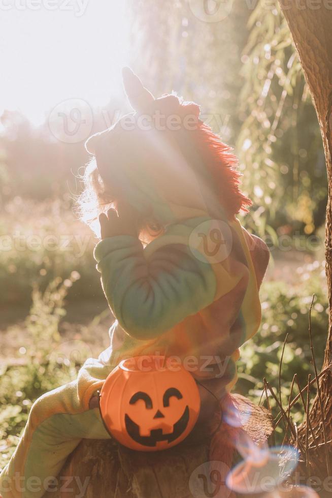 little girl in a rainbow unicorn Halloween costume and a dachshund in a dress with a pumpkin basket for sweets are sitting on stump at forest sunset. fabulous wonderful magical forest. space for text photo
