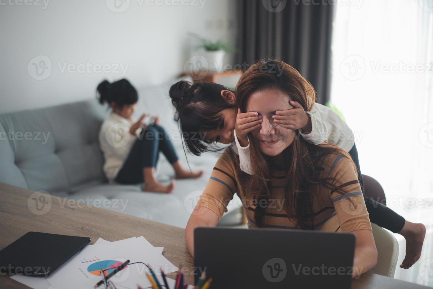 woman sits by the table at home office during lockdown, working on laptop. Playful child distracts from work, covering her mother's eyes, kid making noise and asking attention from busy mom photo