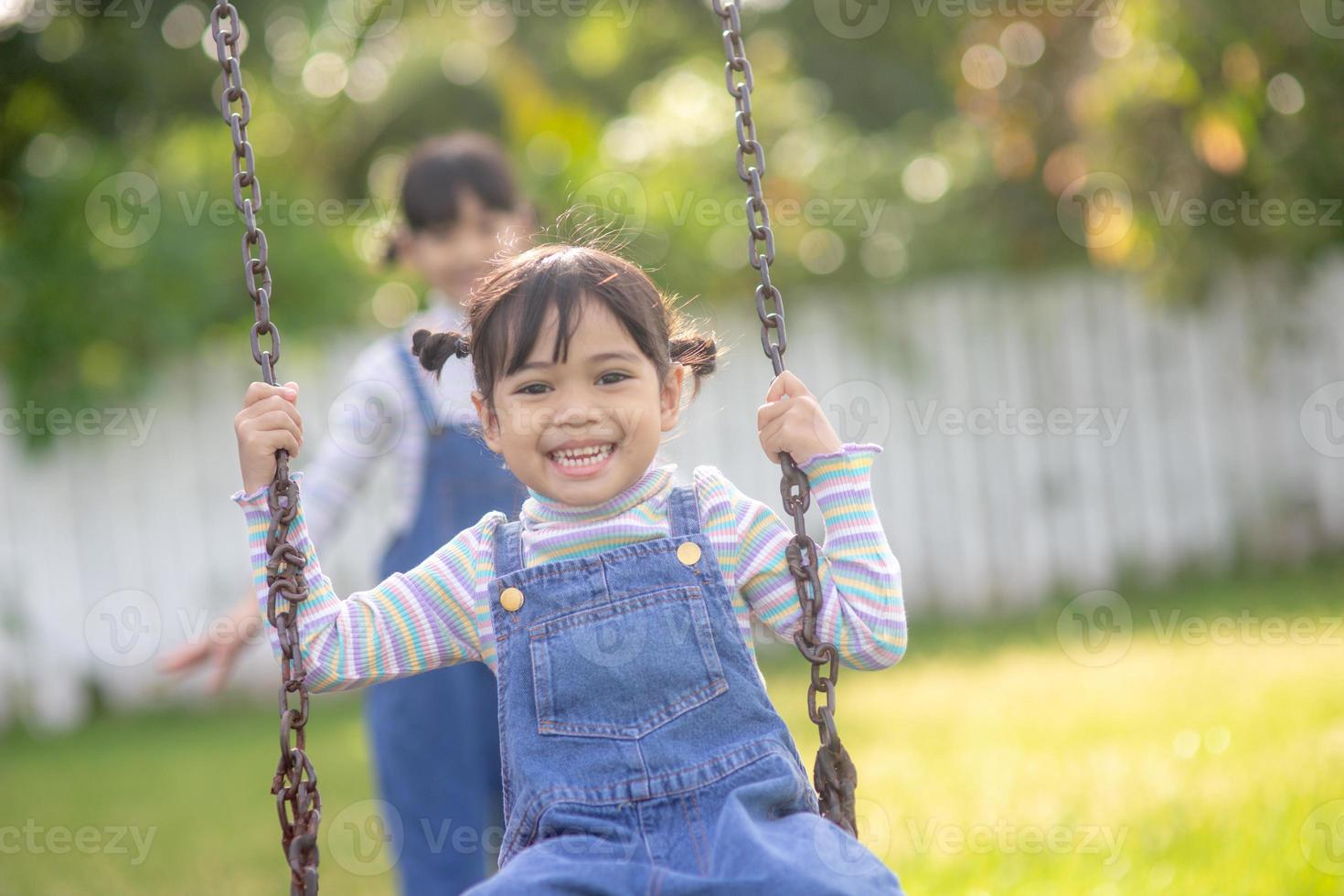 Two cute little sisters having fun on a swing together in a beautiful summer garden on a warm and sunny day outdoors. Active summer leisure for kids. photo