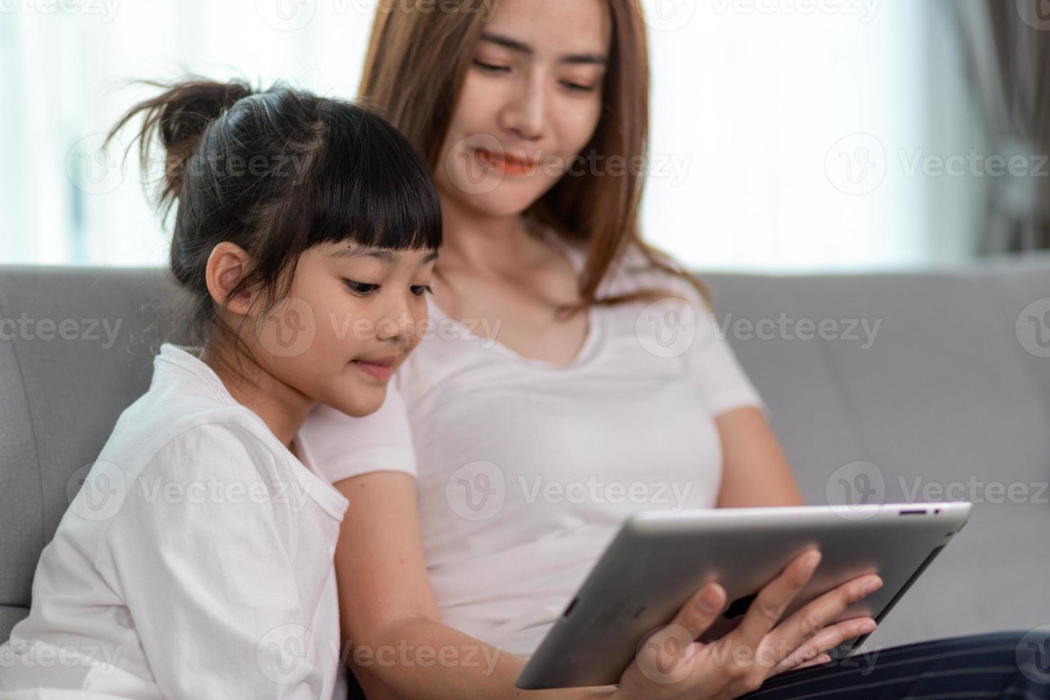 Beautiful Asian mother with daughter sitting on the sofa and playing video game on a tablet while spending time together at home photo