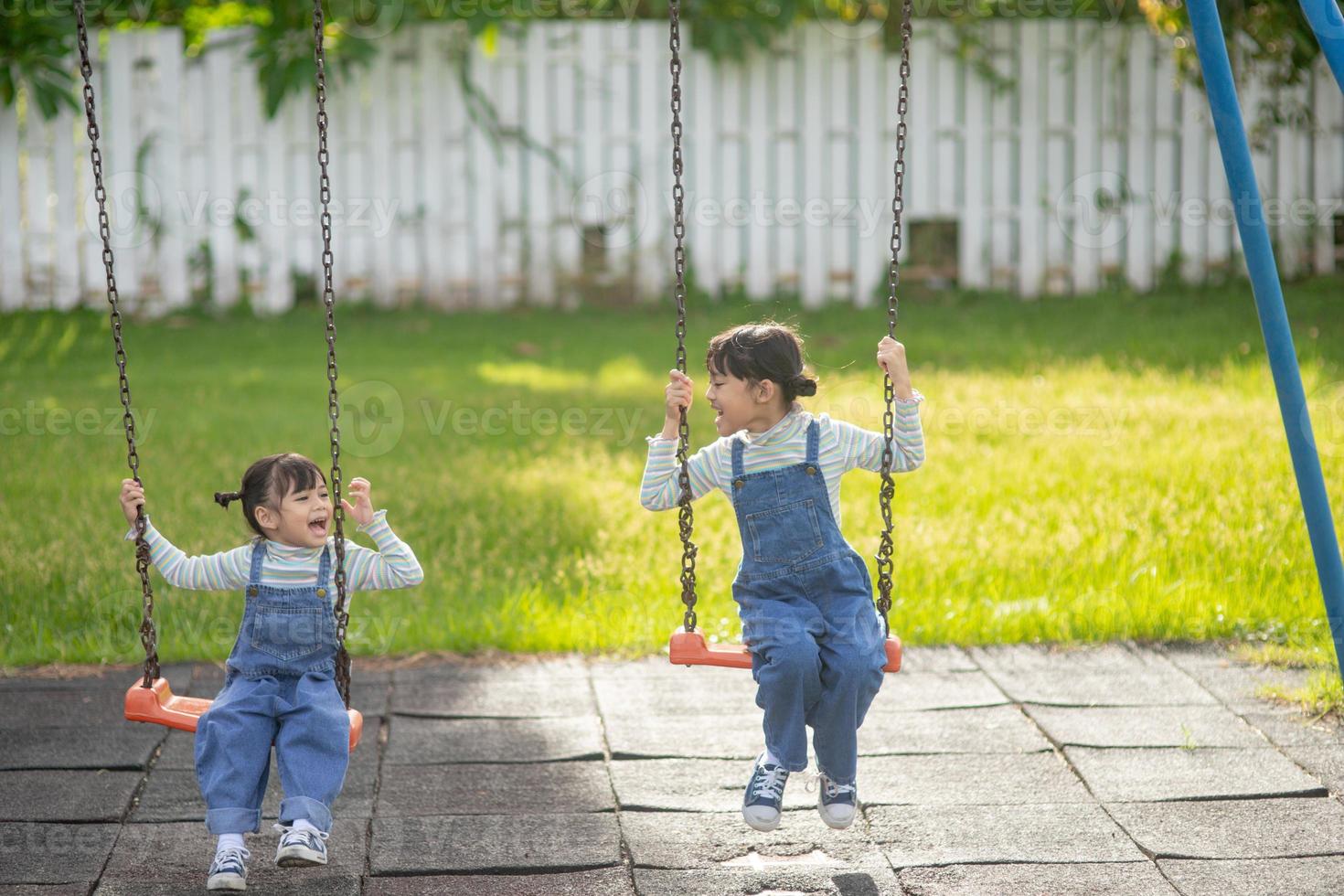 Two cute little sisters having fun on a swing together in a beautiful summer garden on a warm and sunny day outdoors. Active summer leisure for kids. photo