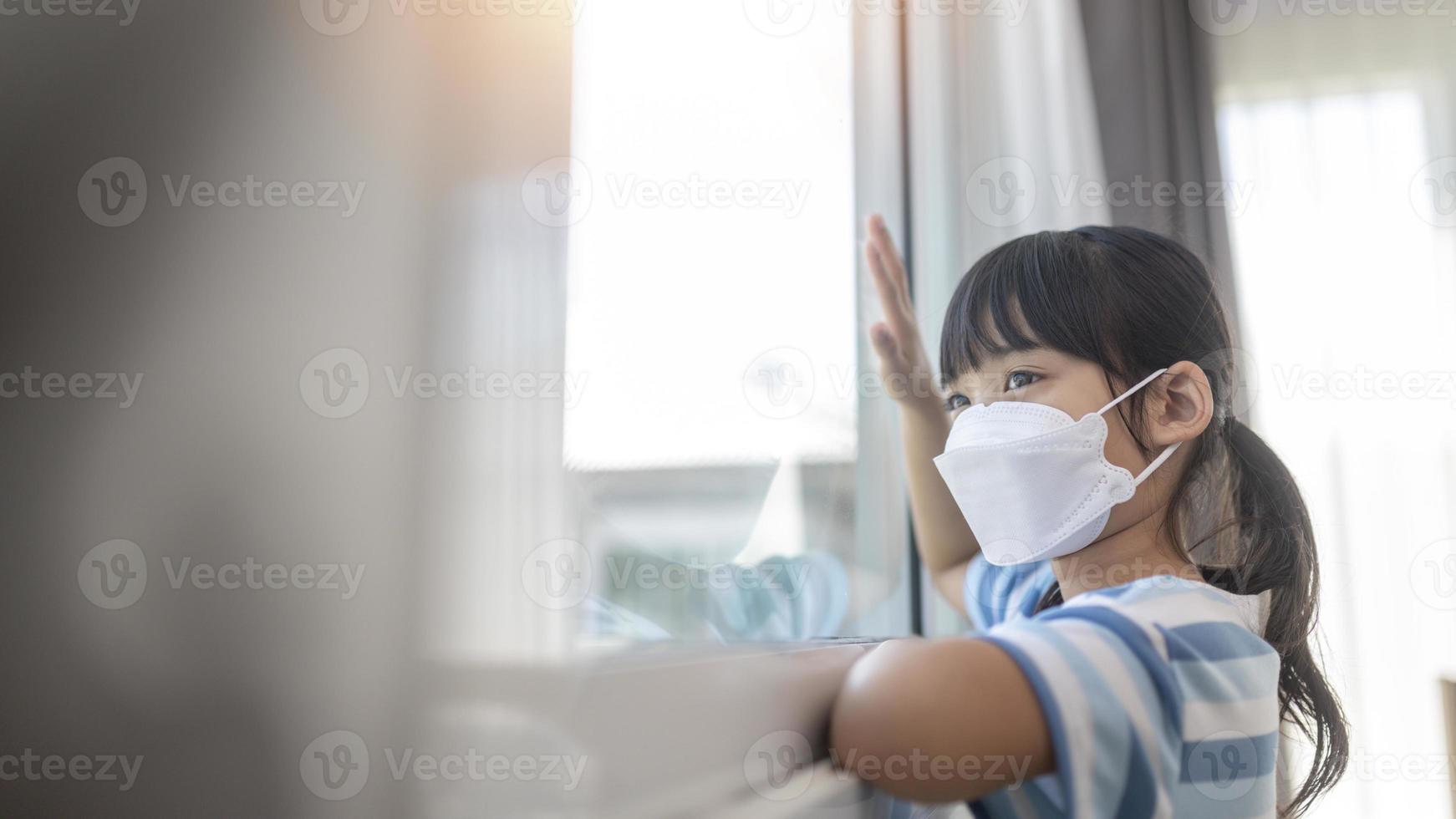 niña asiática con máscara para proteger pm2.5 y covid-19. las niñas enfermas miran la ventana con anhelo en la calle. copie el espacio niño triste en casa aislado foto