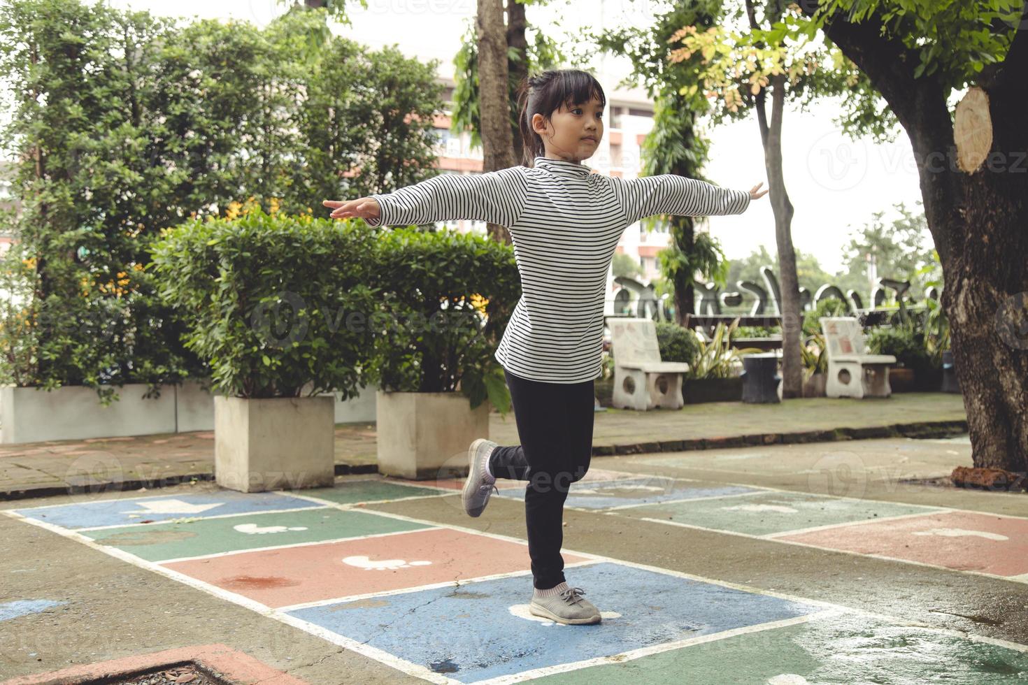 Cute Asian little girl playing hopscotch outdoor. Funny activity game for kids on the playground outside. Summer backyard street sport for children. Happy childhood lifestyle. photo