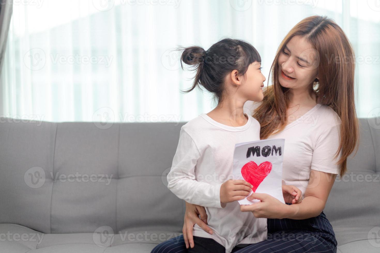 Asian lovely sweet preschool daughter congratulates mommy with life event birthday holiday, prepares for her handmade post card with red painted heart symbol of unconditional love, Mother Day concept photo