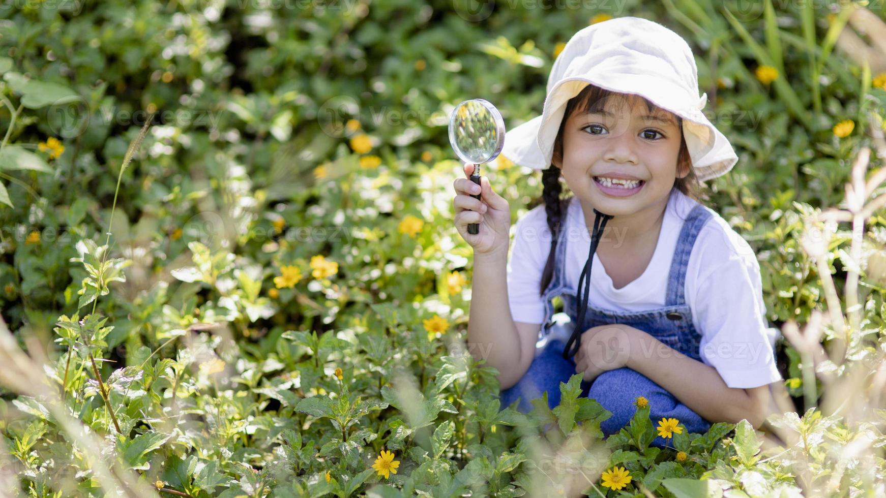 Asian little girl is looking at tree leaves through magnifier, outdoor shoot photo