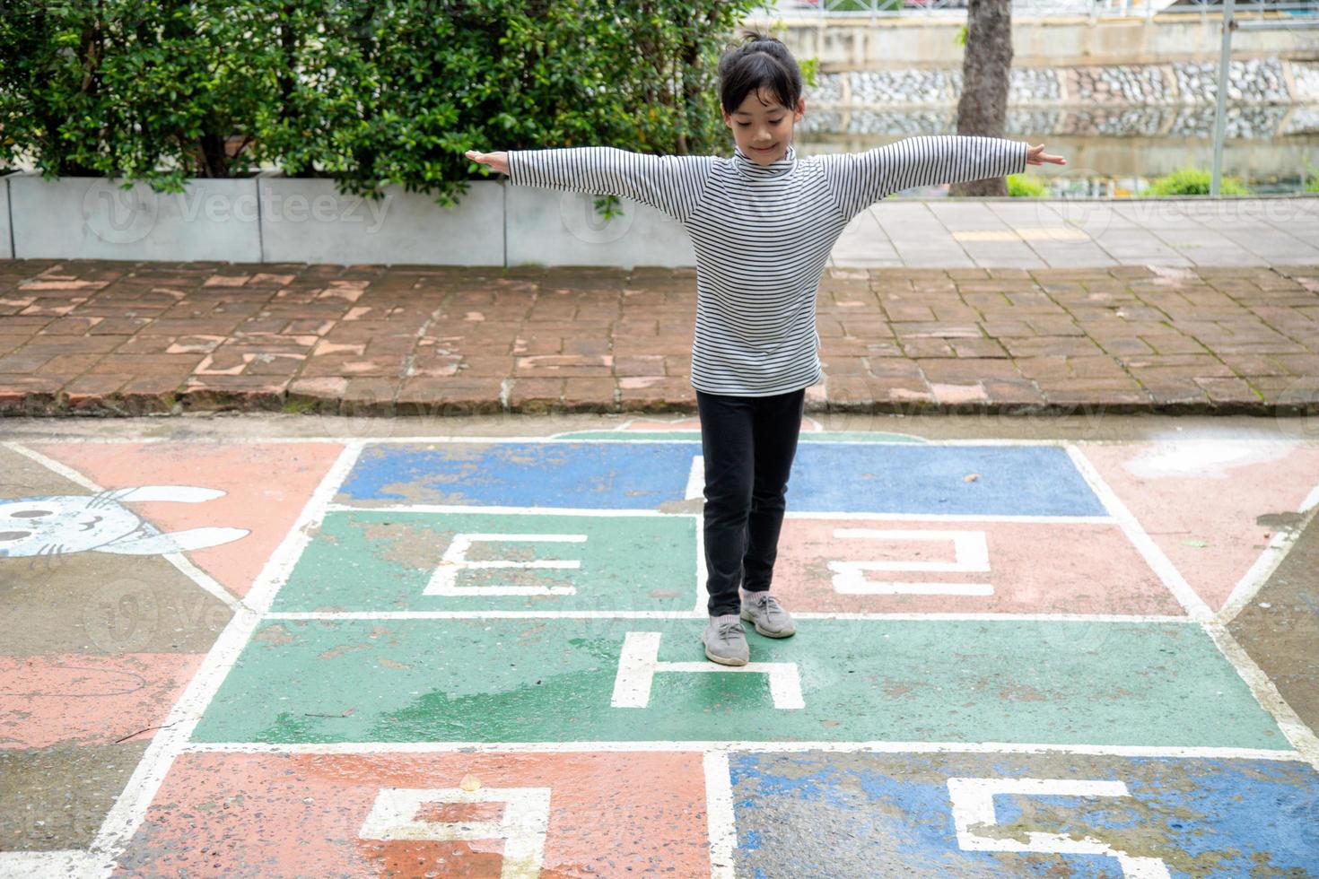 Cute Asian little girl playing hopscotch outdoor. Funny activity game for kids on the playground outside. Summer backyard street sport for children. Happy childhood lifestyle. photo