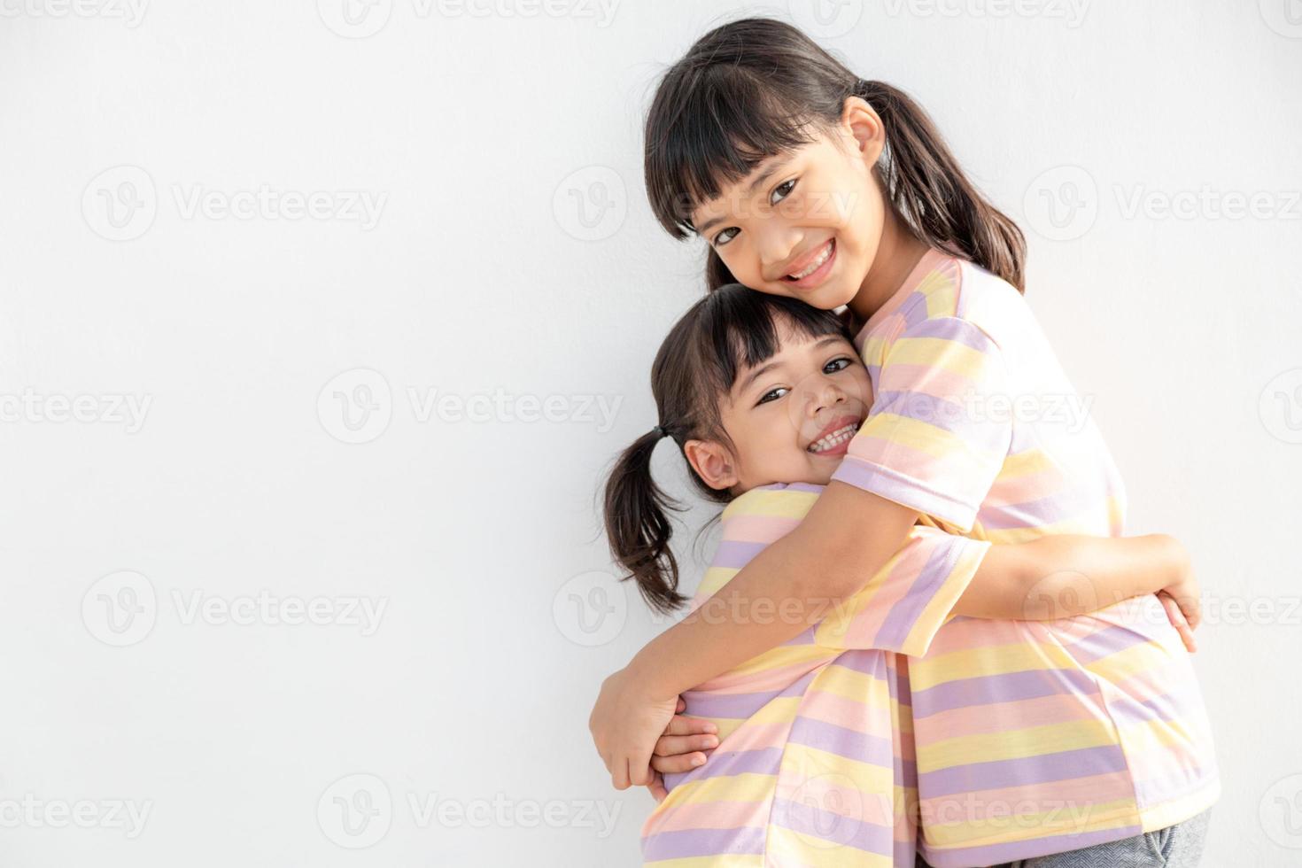 hermanos asiáticos felices y lindos abrazándose, sintiendo amor y conexión, hermana niña sonriente abrazando a su hermana pequeña de fondo blanco, 2 niños con buenas relaciones foto