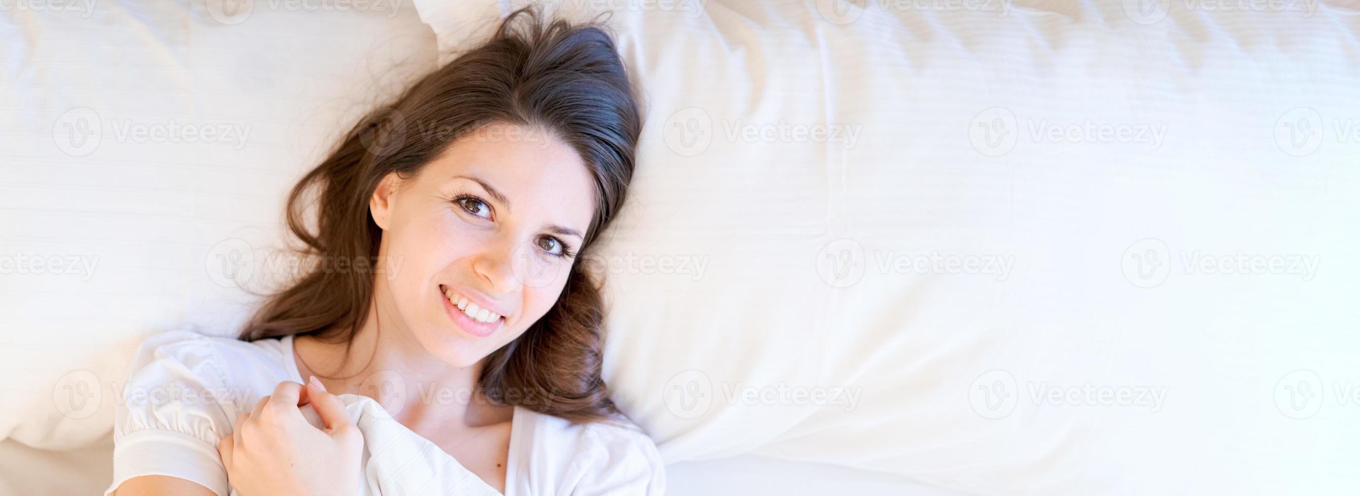 Dreamy girl lying on white bed with large pillows, hugging blanket with her photo