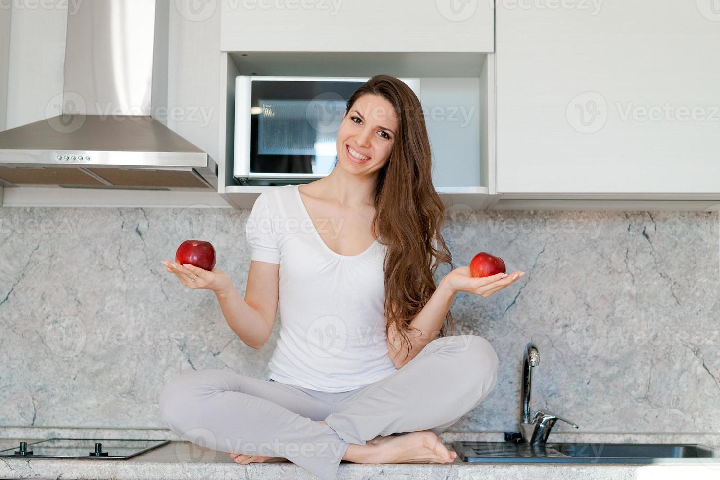 Young slim woman standing in kitchen on table, holding red apples, looking photo