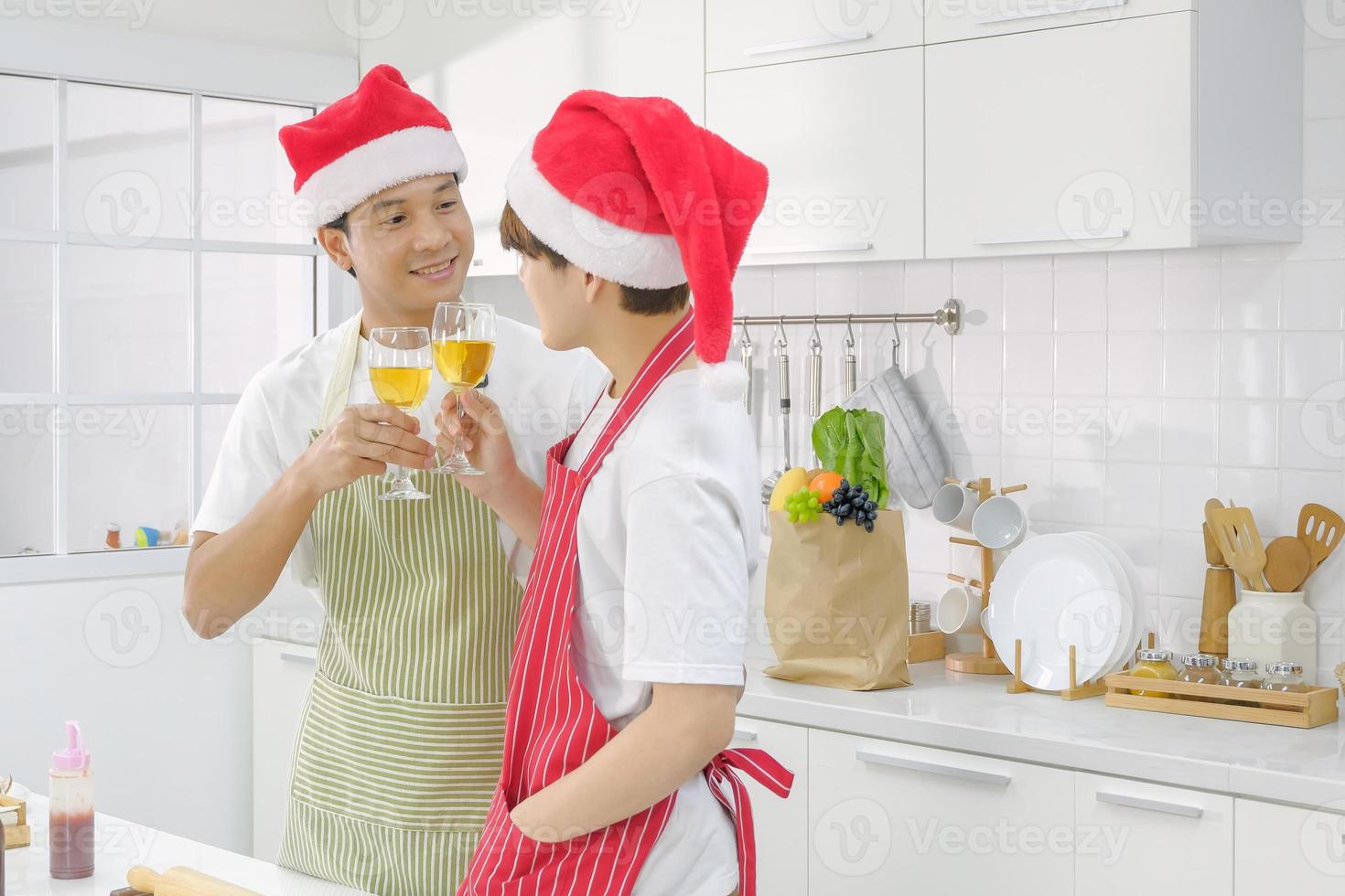 joven pareja lgbt concepto de vida doméstica. feliz pareja de jóvenes asiáticos con sombrero de santa y copa de vino tintineante para celebrar la fiesta privada de año nuevo de navidad en la cocina blanca. enfoque selectivo. foto