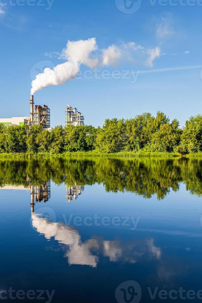 pipes of woodworking enterprise plant sawmill with beautiful reflection in blue water of river. Air pollution concept. Industrial landscape environmental pollution waste of thermal power plant photo