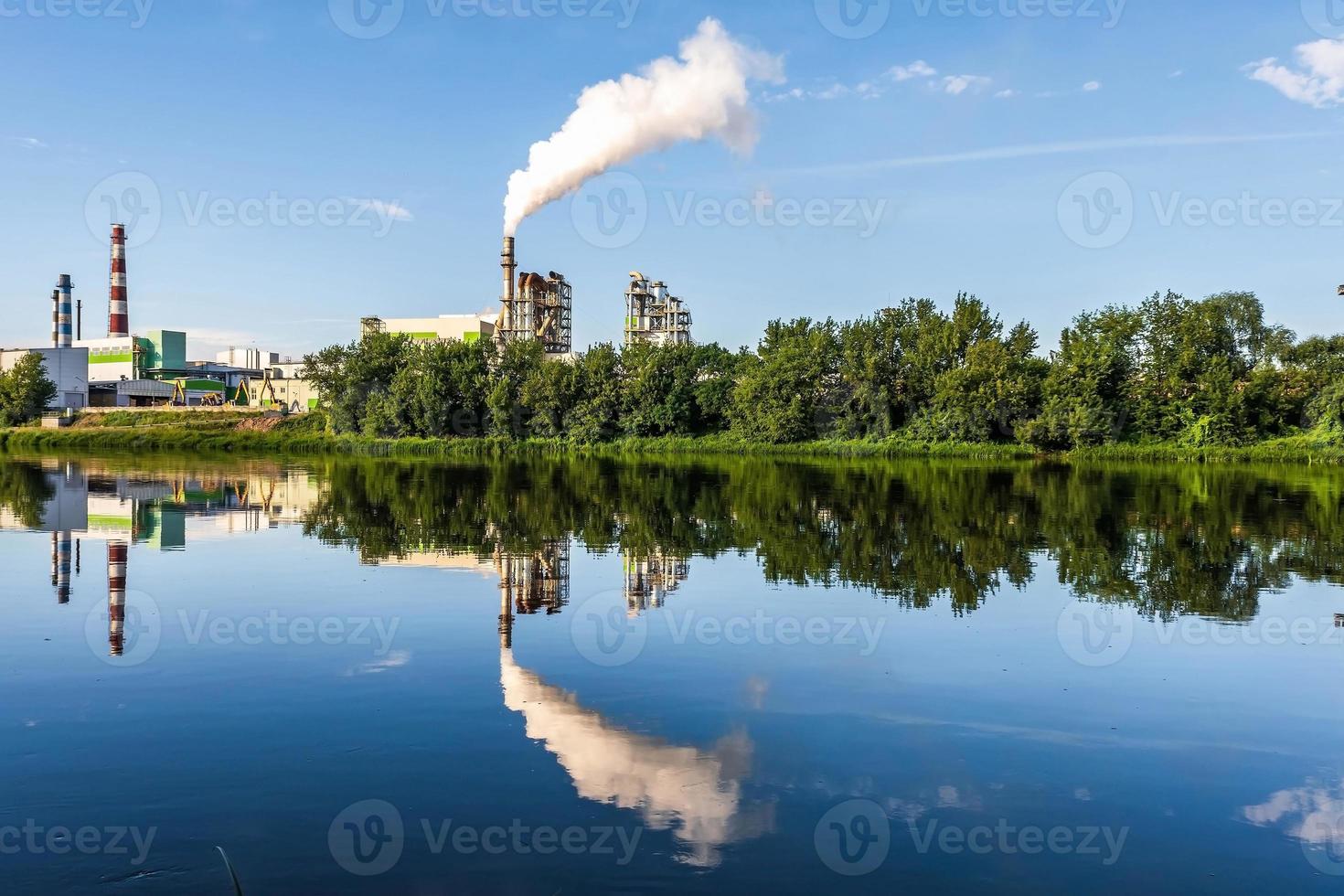 tuberías de aserradero de planta empresarial de carpintería con un hermoso reflejo en el agua azul del río. concepto de contaminación del aire. paisaje industrial contaminación ambiental residuos de central térmica foto