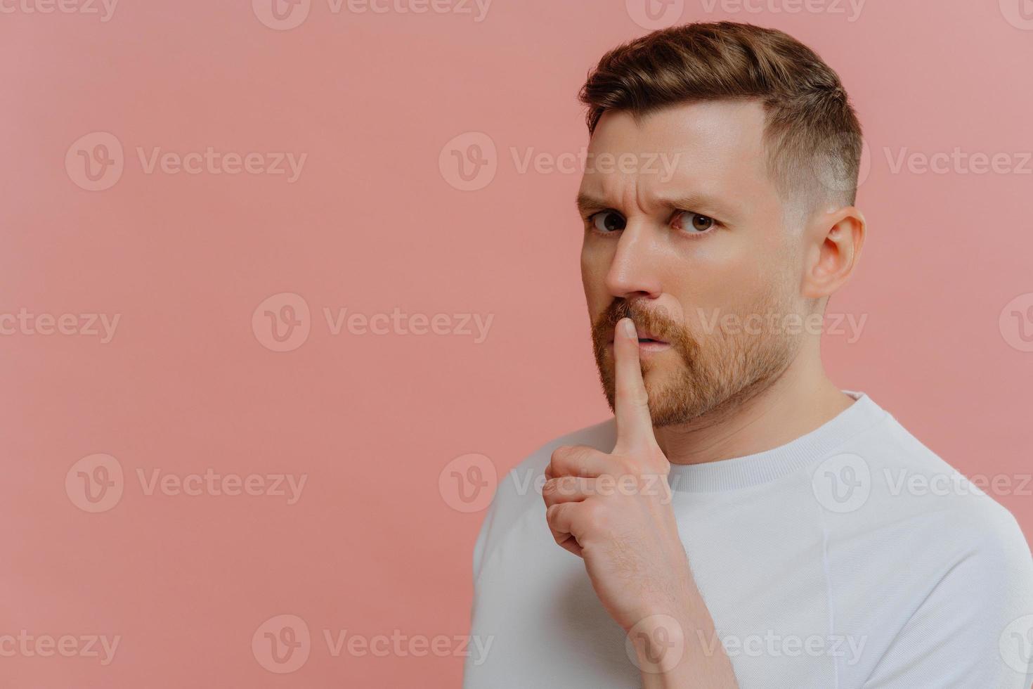 Handsome serious man makes silence gesture presses index finger to lips spreads rumors asks to be quiet looks with strict expression dressed in casual white t shirt isolated over pink background photo