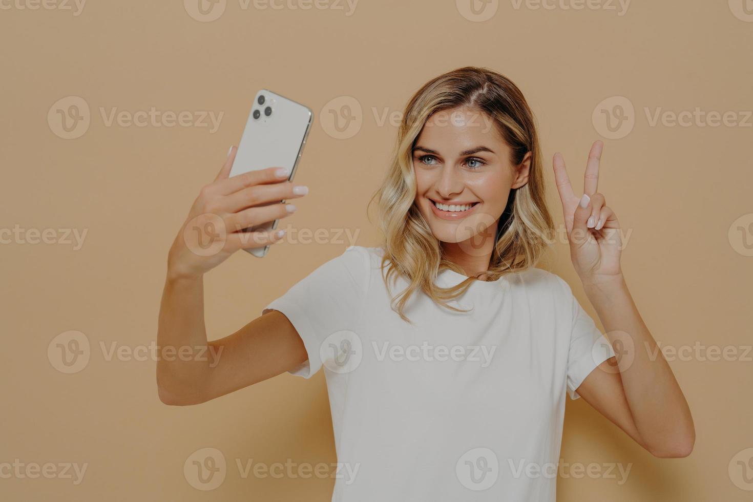 Cool joyful woman with blonde hair with smartphone in hand making selfie in studio against nude background photo