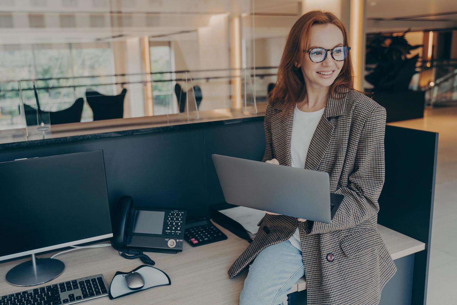 Smiling female office employee sitting on desk at her workplace while holding laptop photo