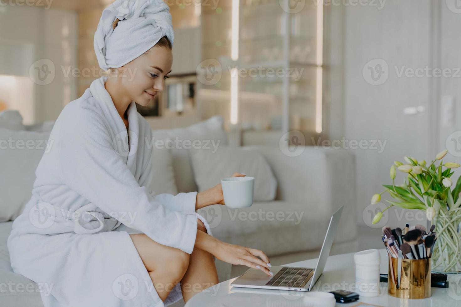Indoor shot of female freelancer workds distantly rom home types on laptop computer dressed in bathrobe drinks coffee or tea enjoys domestic atmosphere surfs internet reads advice about skin care photo