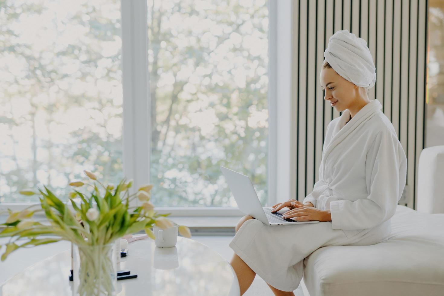 Sideways shot of attractive refreshed Caucasian woman sits at comfortbale sofa works on laptop computer wears bathrobe and wrapped towel on head poses against big window in cozy spacious room photo