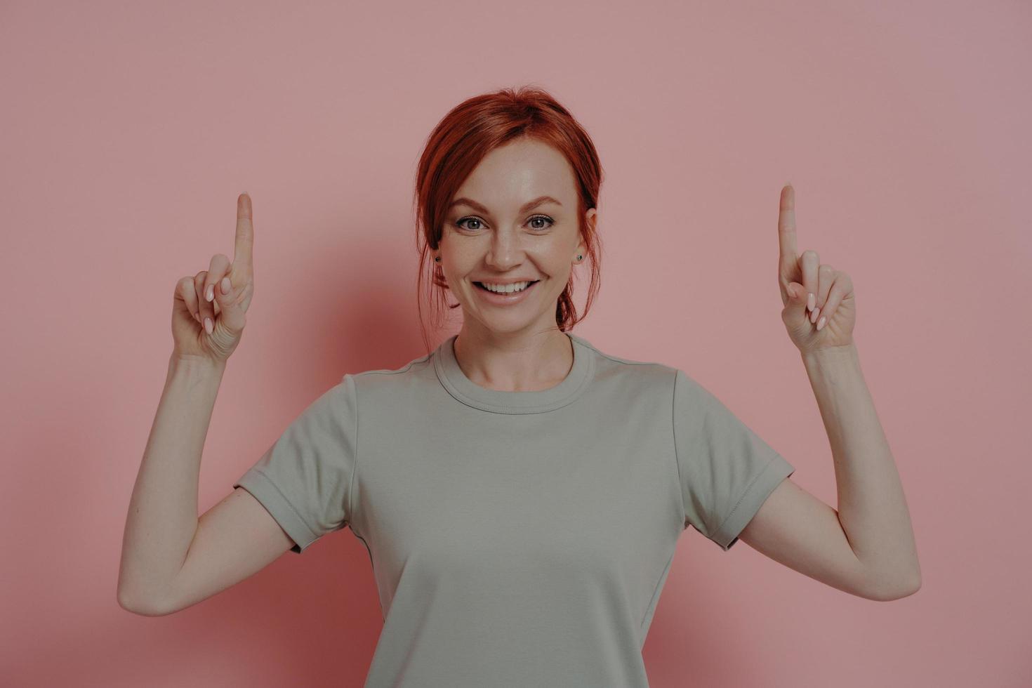 Happy young caucasian woman pointing upwards with index fingers and smiling cheerfully photo