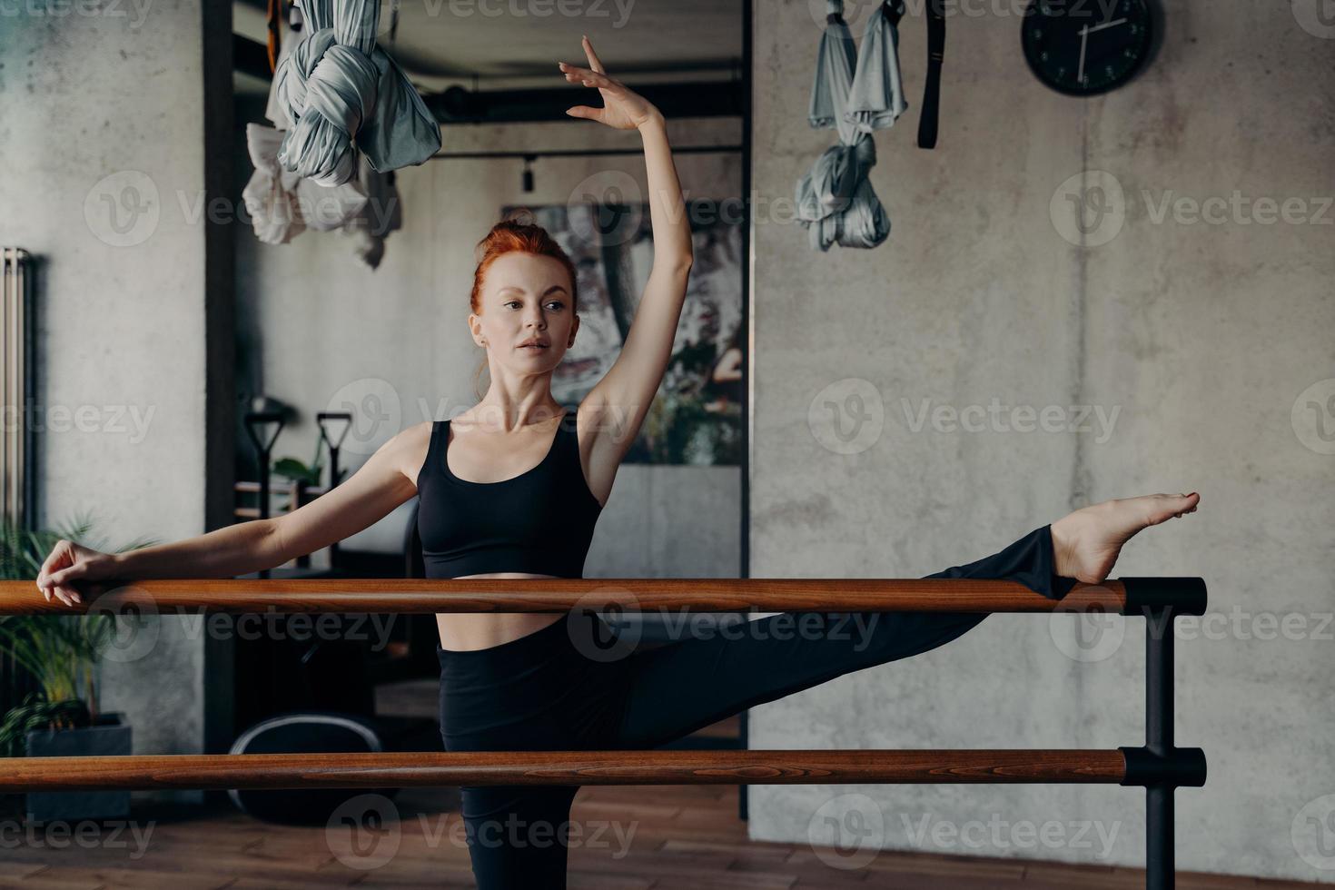 joven mujer atlética de jengibre estirando la pierna en la barra de ballet con una mano levantada durante el entrenamiento de la barra foto