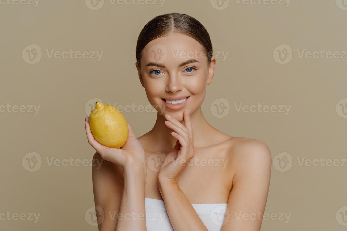 Cosmetology skin care and beauty concept. Pleased brunette young woman wrapped in bath towel holds lemon for making facial mask has refreshed clean skin after taking shower and spa procedures photo