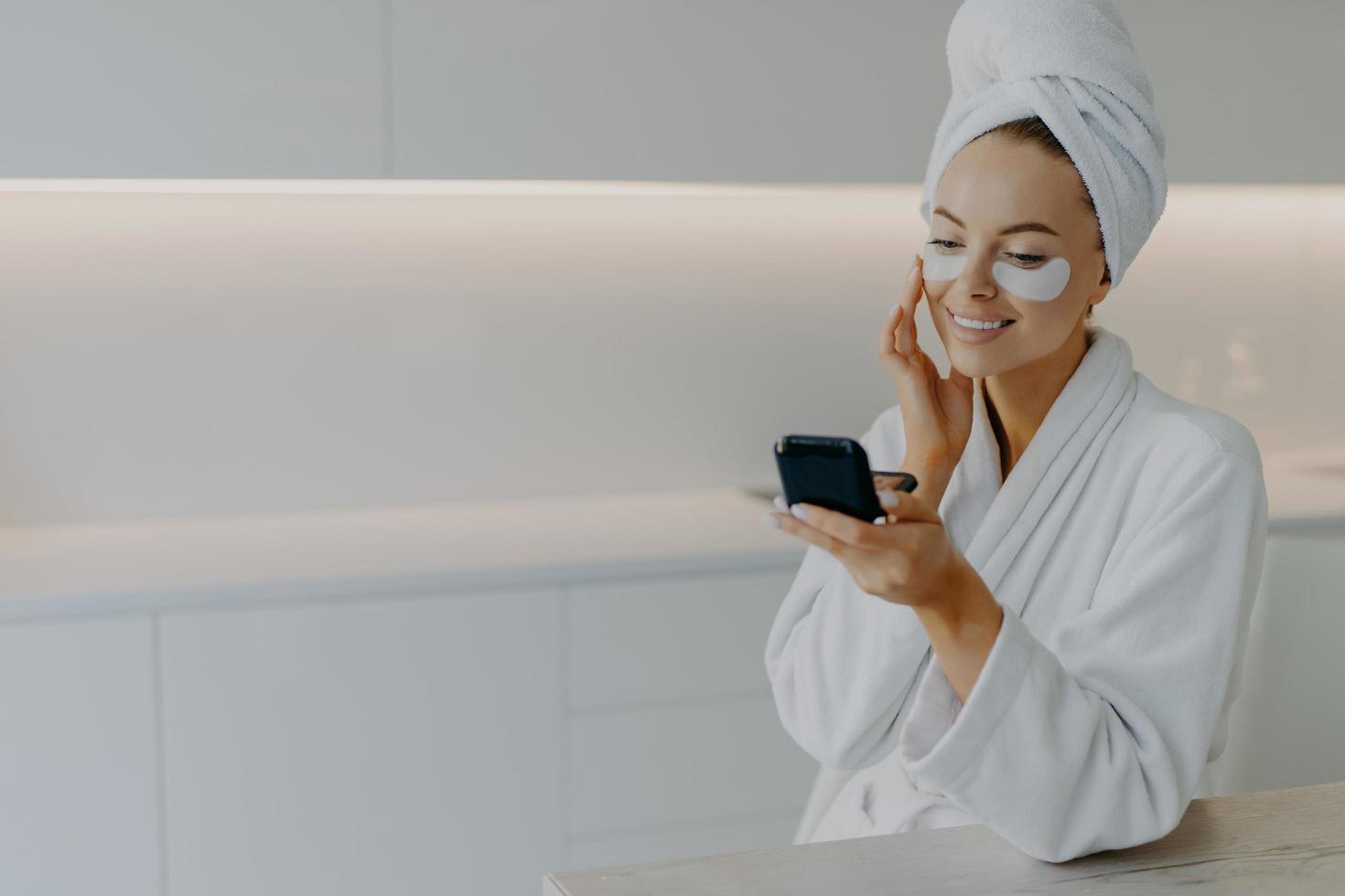 Beautiful happy young lady applies collagen patches under eyes looks at herself in mirror does rejuvenation procedure wears bath towel and dressing gown after taking shower has skin care routine photo