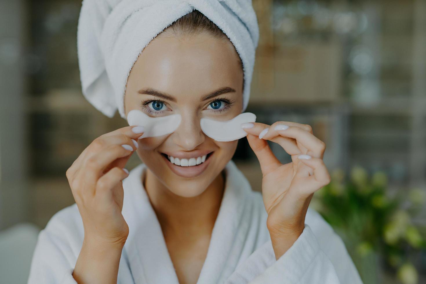 una foto de una joven feliz de ojos azules con un mínimo de maquillaje se quita los parches de belleza disfruta de un buen efecto tiene una piel sana y bien cuidada dientes blancos perfectos usa una bata de baño y una toalla envuelta en la cabeza