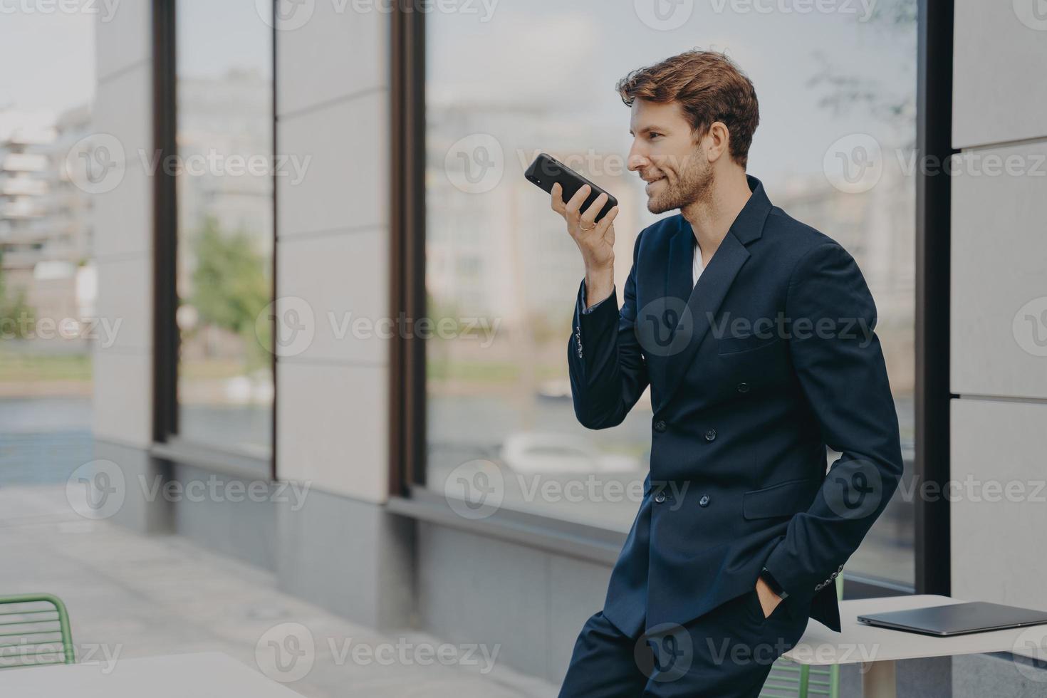 Confident businessman with smartphone using voice assistant app while leaning on cafe table outdoors photo