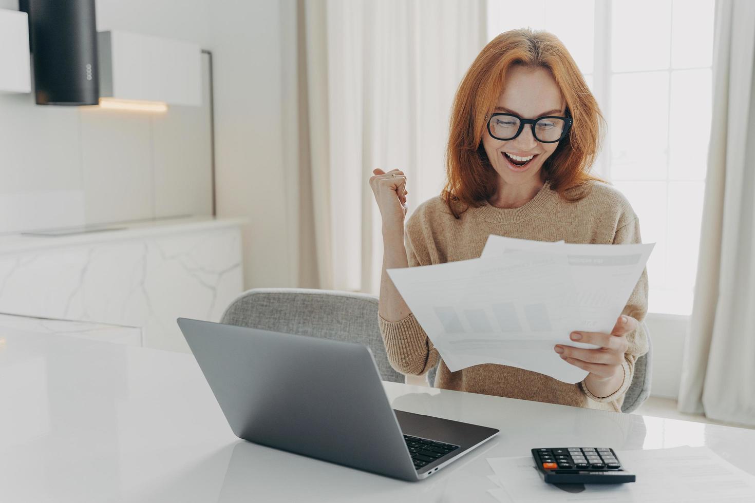 Female freelancer checks calculations clenches fist looks happily at paper documents photo
