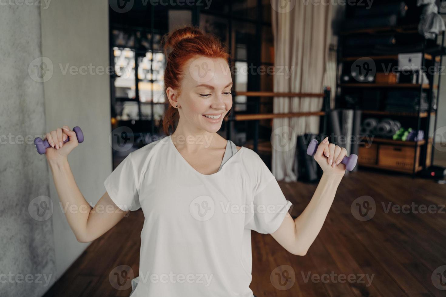 Smiling sportive redhead woman exercising with dumbbells in fitness studio photo