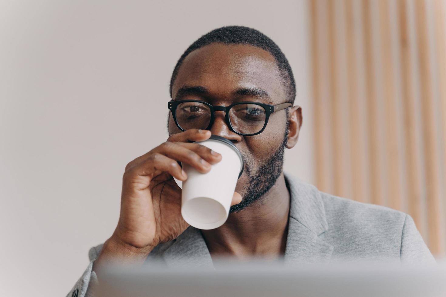 Attractive confident Aframerican man company CEO drinking coffee while looking at laptop screen photo