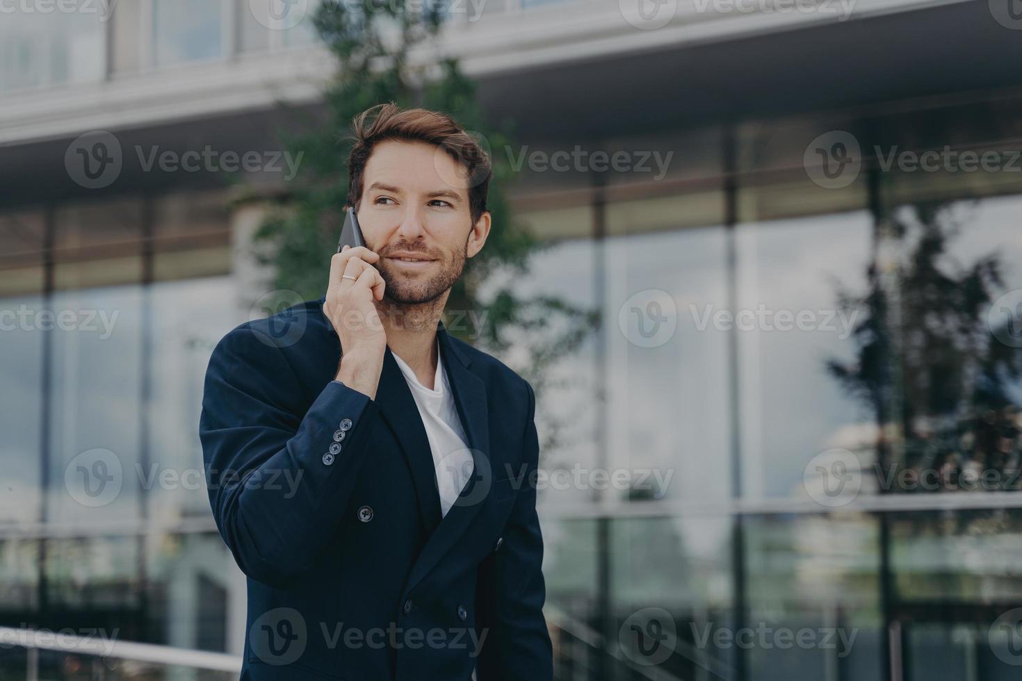 Male entrepreneur talks via phone solves urgent problems poses near business center photo