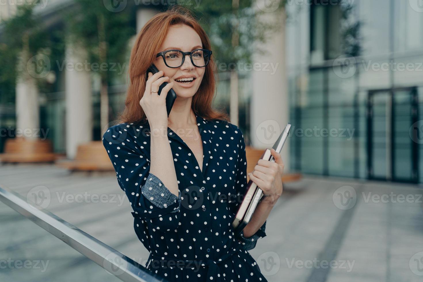 Cheerful redhead European woman talks on modern mobile phone has glad expression photo