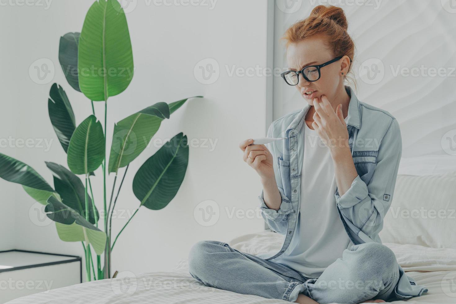 Worried ginger young woman checks pregnancy result test finds out about future maternity sits on bed photo