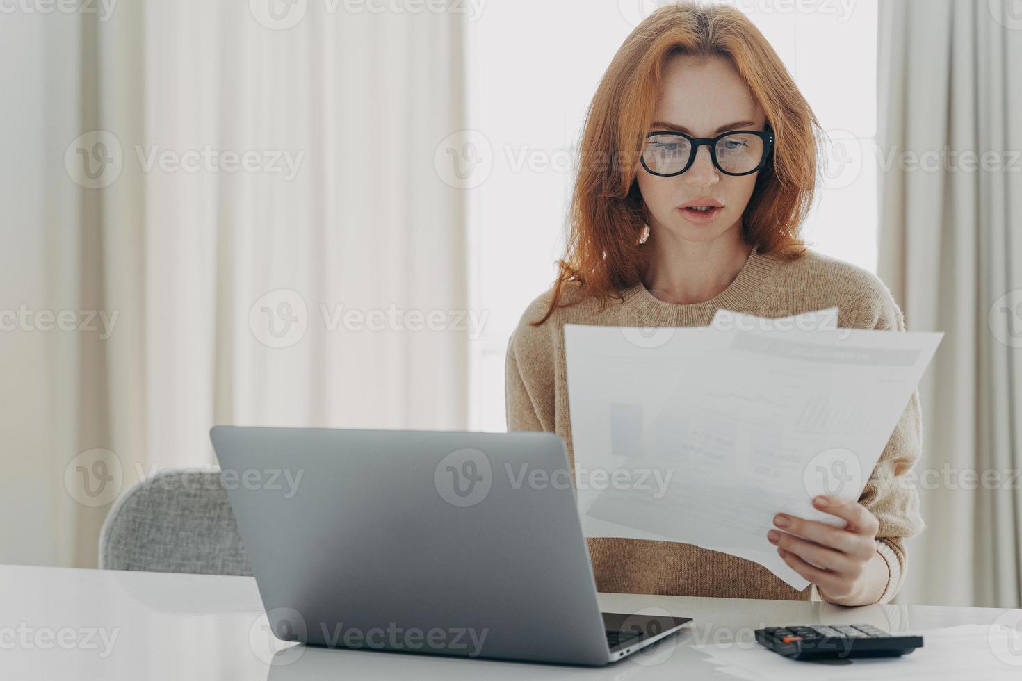 Serious redhead Caucasian woman calculates bills holds paper documents has focused look photo