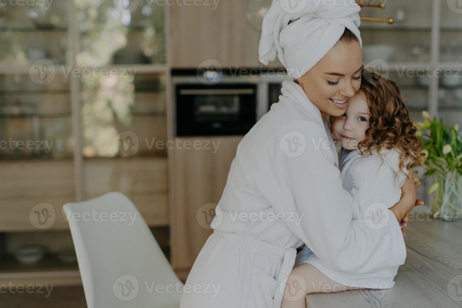 Happy affectionate young mother in bath towel and dressing gown embraces with love who sits at table feel refreshed after taking shower spend free time at home. Family beauty wellness concept photo
