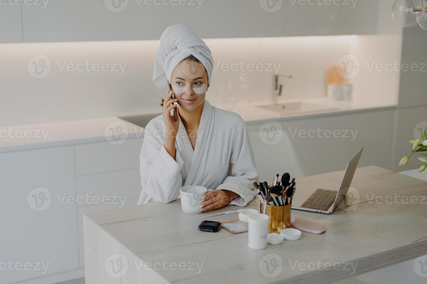 Lovely female model wears bathrobe and soft towel wrapped on head undergoes skin care procedures applies beauty patches under eyes has coffee break while talking with friend via cellular being at home photo