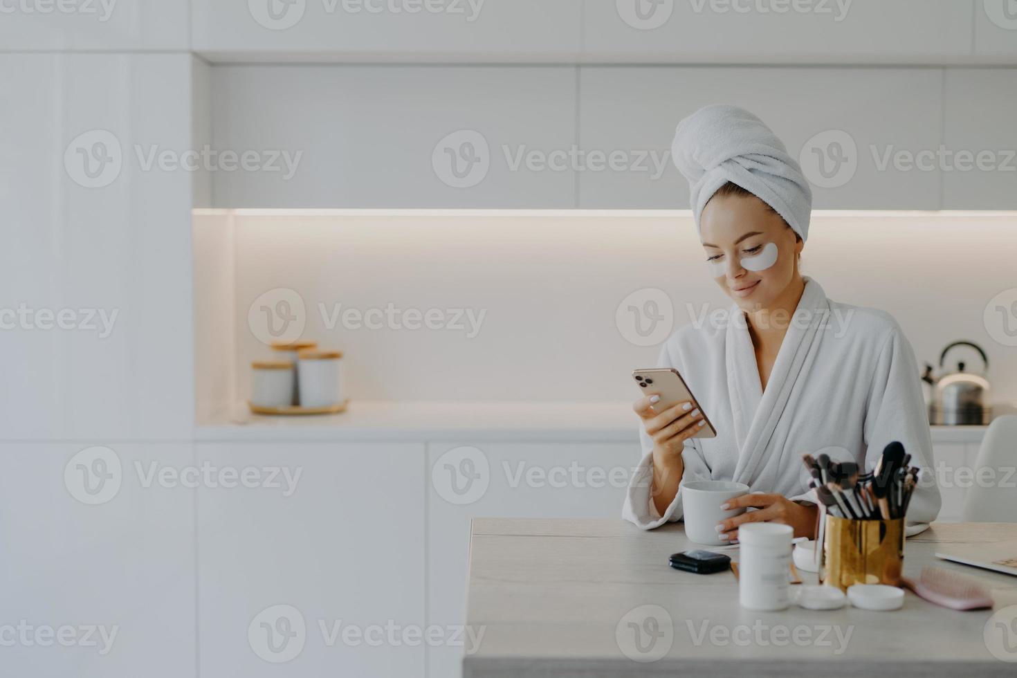 Glad young European woman dressed in bathrobe types text messages in online chat during coffee break undergoes skin care procedures applies beauty patches poses over kitchen interior at home photo