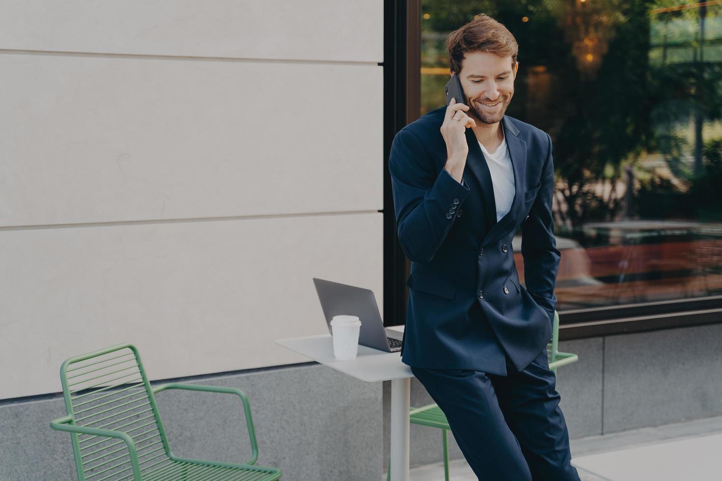 exitoso comerciante masculino guapo discutiendo los precios de las acciones mientras se apoya en la mesa de café de la acera foto