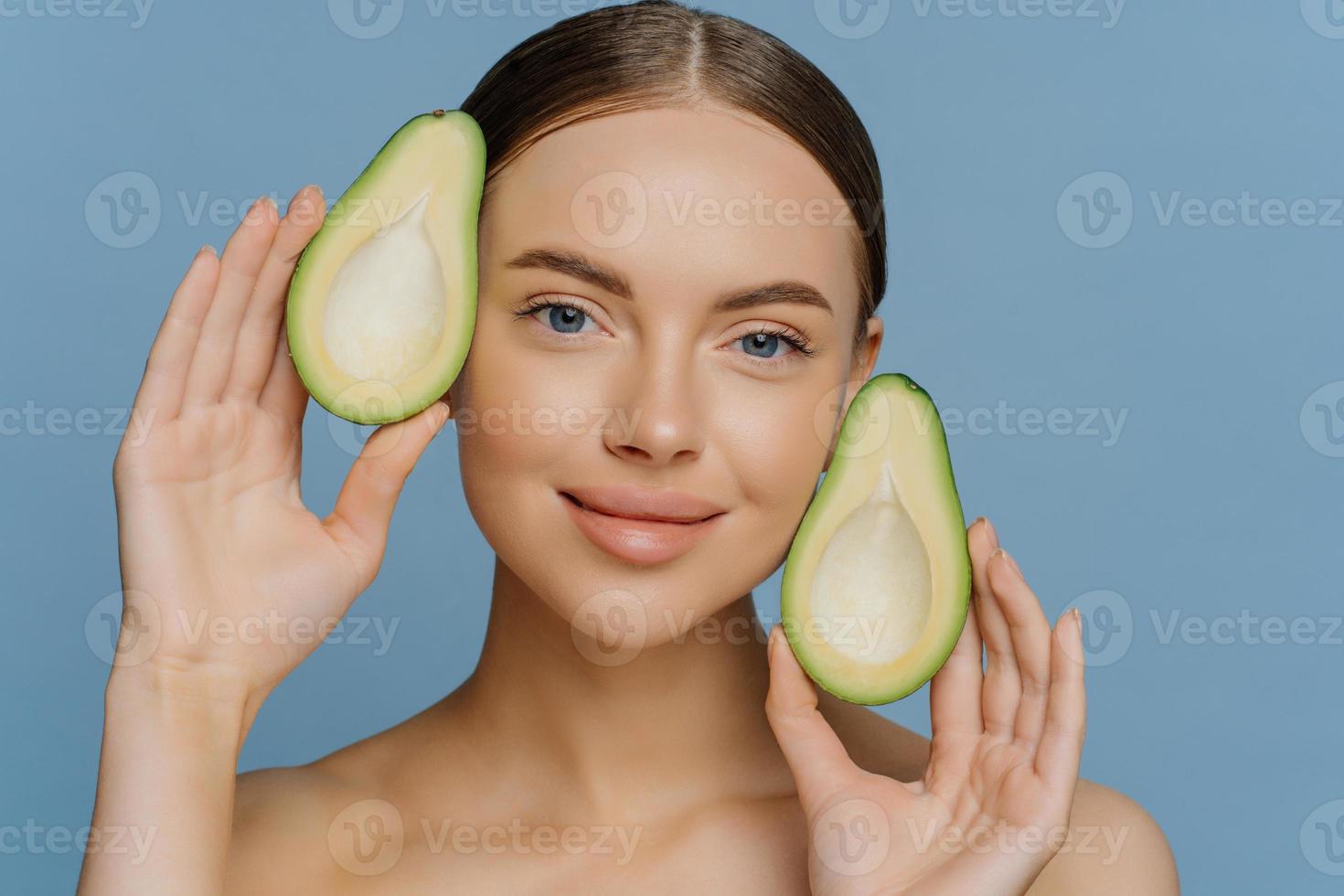 tratamientos de belleza para el cuidado de la piel. una joven modelo europea morena complacida sostiene mitades de aguacate cerca de las poses de la cara con el cuerpo desnudo aislado sobre fondo azul. grasas saludables. cosméticos ecológicos foto