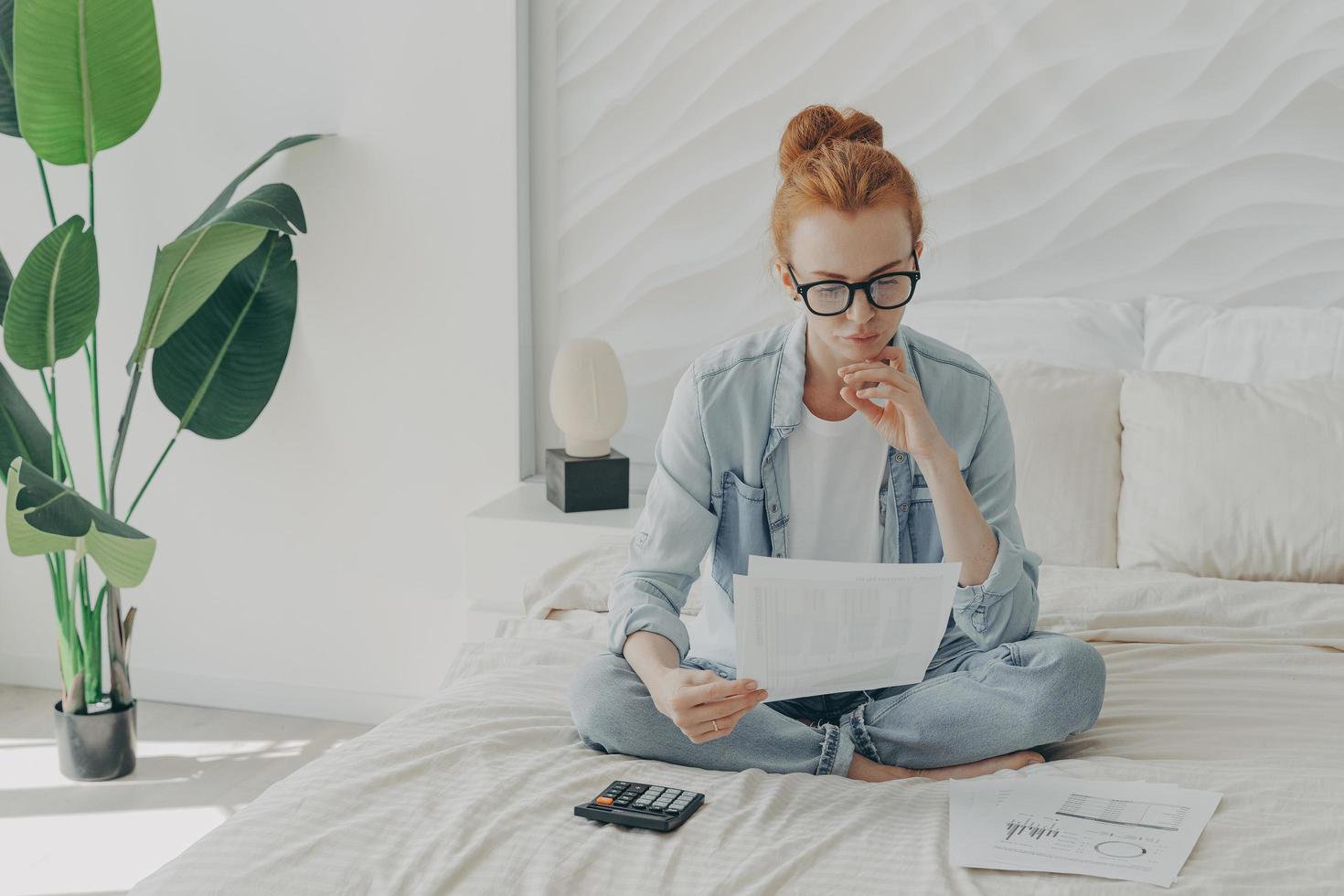 Focused ginger european woman sitting on bed and calculating domestic bills at home, holding papers photo