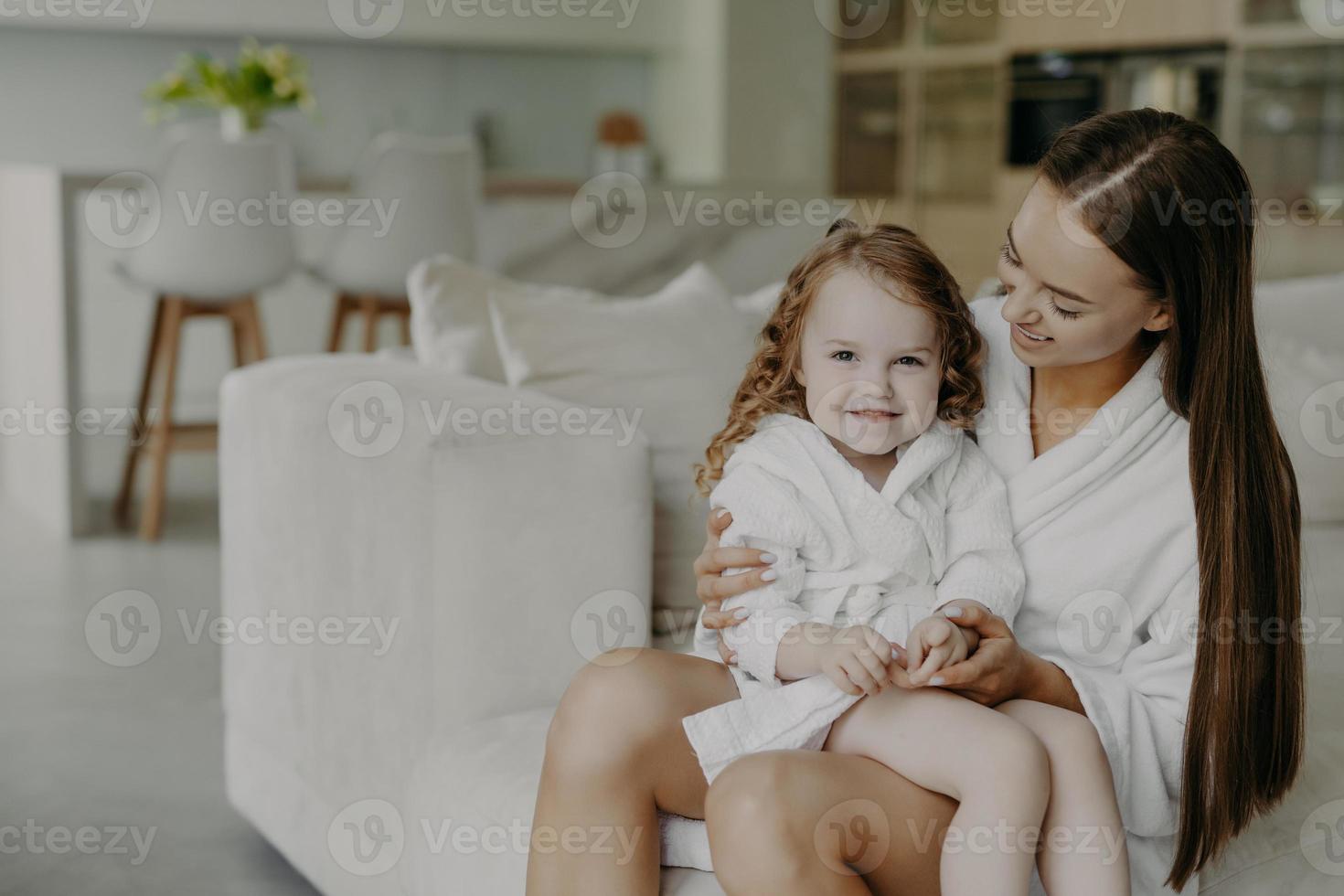 Beautiful brunette affectionate mother holds small curly haired cute dauhter in dressing gown pose in cozy room against modern apartment interior. Mom and little girl at home after taking shower photo