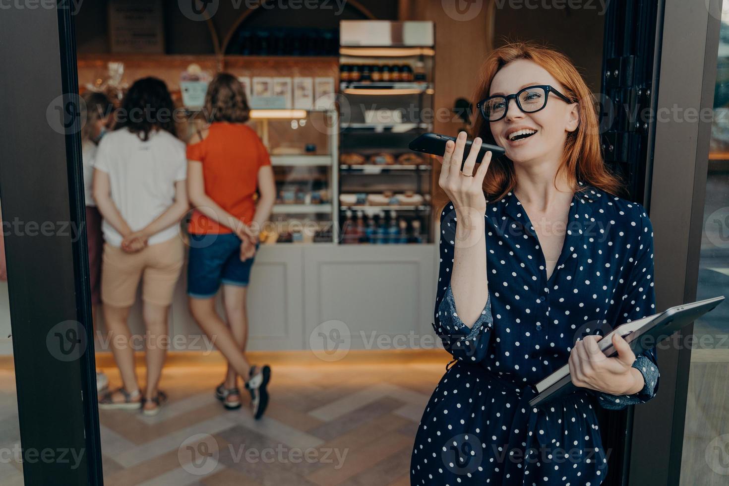 Redhead young woman makes voice call keeps smartphone near mouth wears spectacles polka dot dress photo