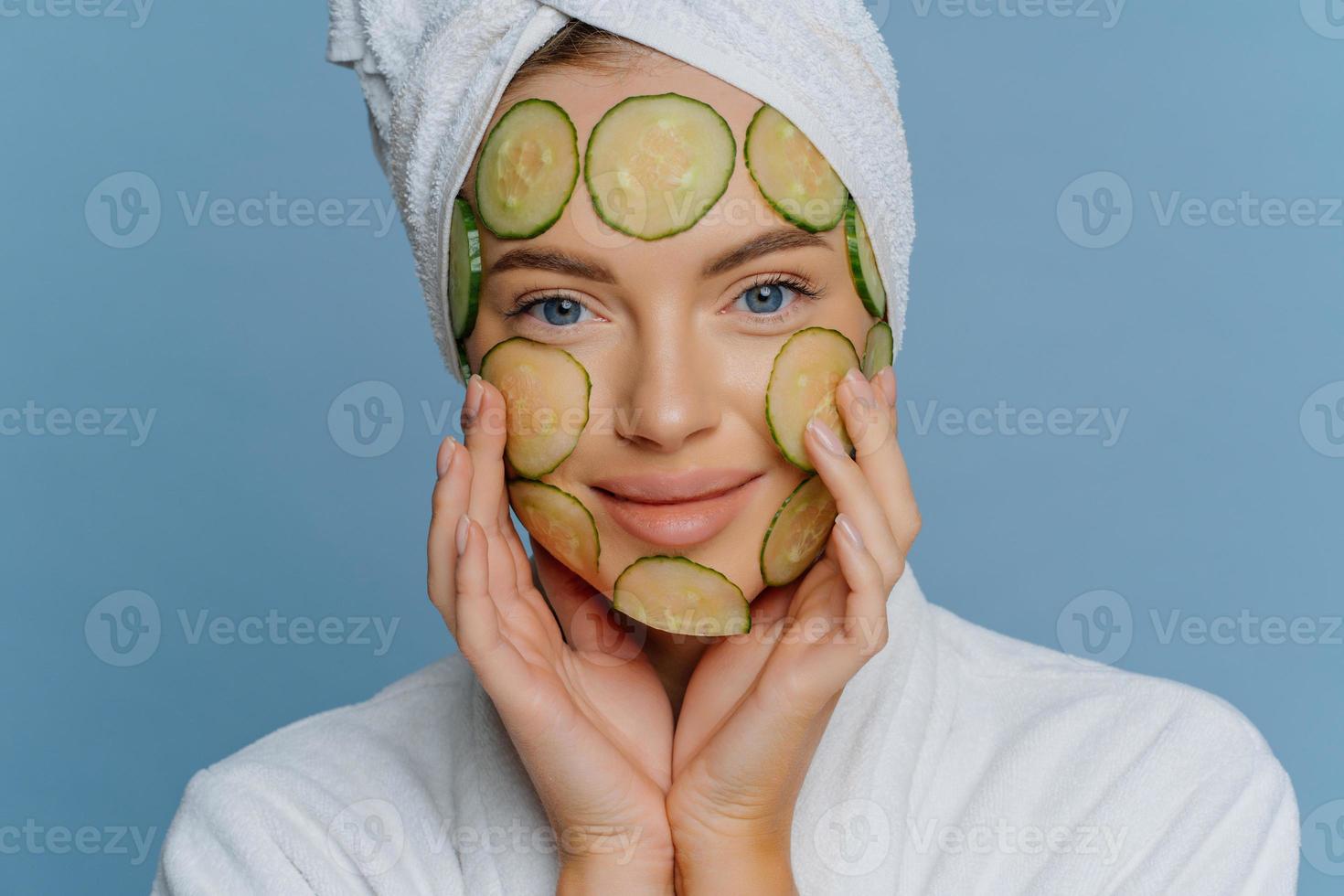 Close up shot of young woman applies cucumber slices on face for healthy skin looks directly at camera dressed in bath dressing gown and towel on head isolated on blue background. Facial care concept photo