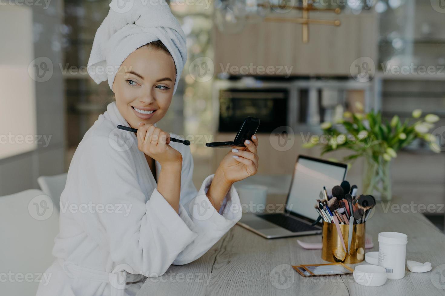 Indoor shot of smiling woman applies makeup after taking shower holds cosmetic brush and mirror wears bathrobe and wrapped towel on head. Women grooming wellness and beauty treatment concept photo