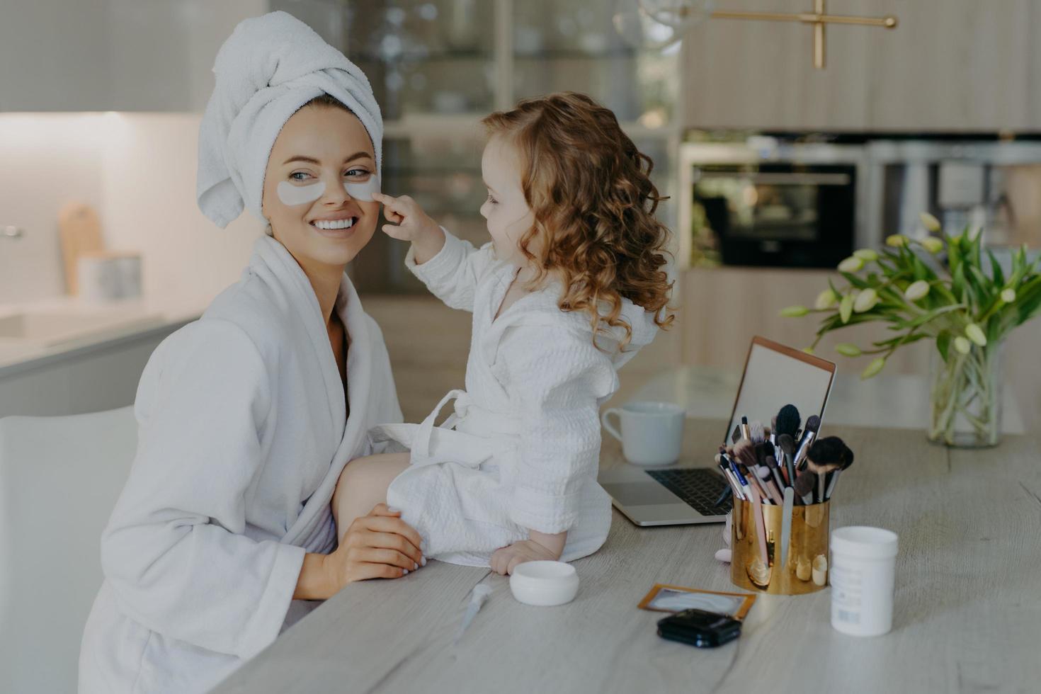 una niña pequeña y bonita con el pelo rizado mira cómo la madre hace los procedimientos cosméticos aplica parches de colágeno debajo de los ojos para el tratamiento de la piel usa albornoces posan cerca del escritorio con productos de belleza. cuidado diario foto