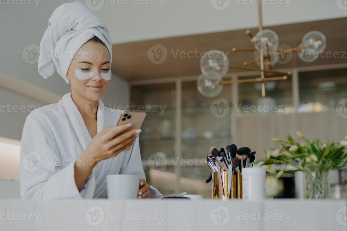 foto de una mujer joven atractiva que usa una toalla de baño en la cabeza y una bata blanca suave sostiene el teléfono móvil disfruta de la comunicación en línea aplica parches humectantes debajo de los ojos bebe bebidas calientes
