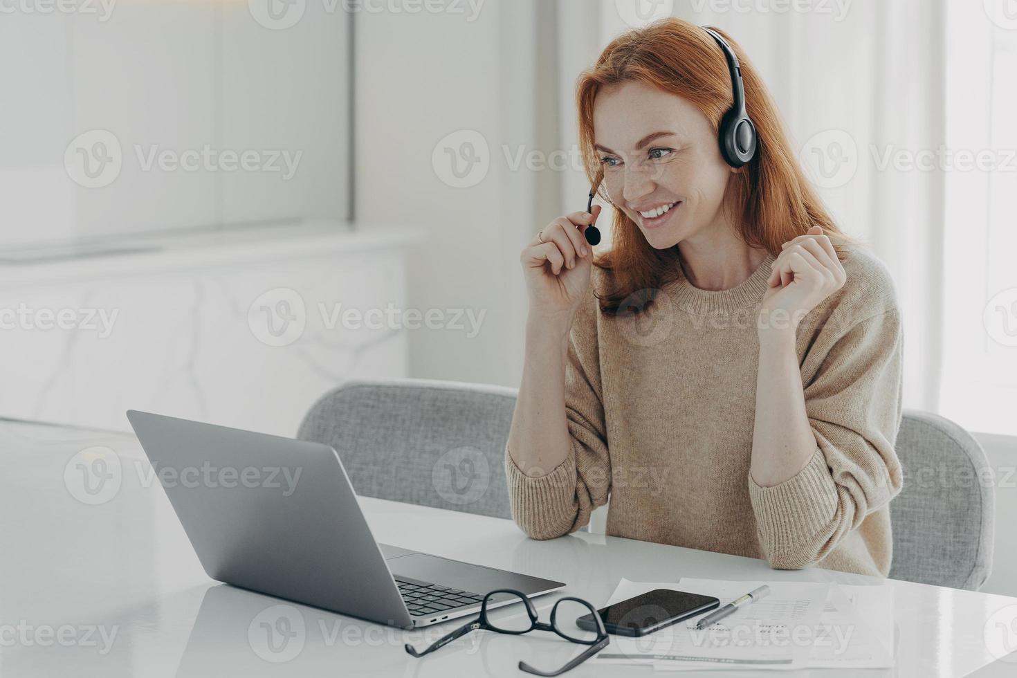 Pleasant smiling redhead woman in headset communicating with colleagues during video call on laptop photo