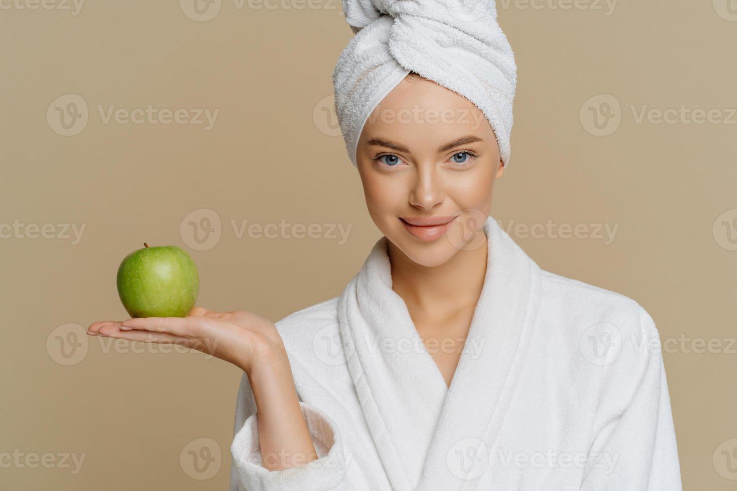 Studio shot of young woman moisturizes face with fresh apple dressed in shite coat wrapped towel on head after taking shower isolatedover brown background. Natural beauty and hygiene concept photo